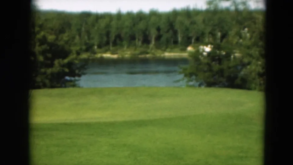 1975 Duluth 8mm frame showing a green golf course near a calm lake and dense trees, with no clear person in this quadrant.