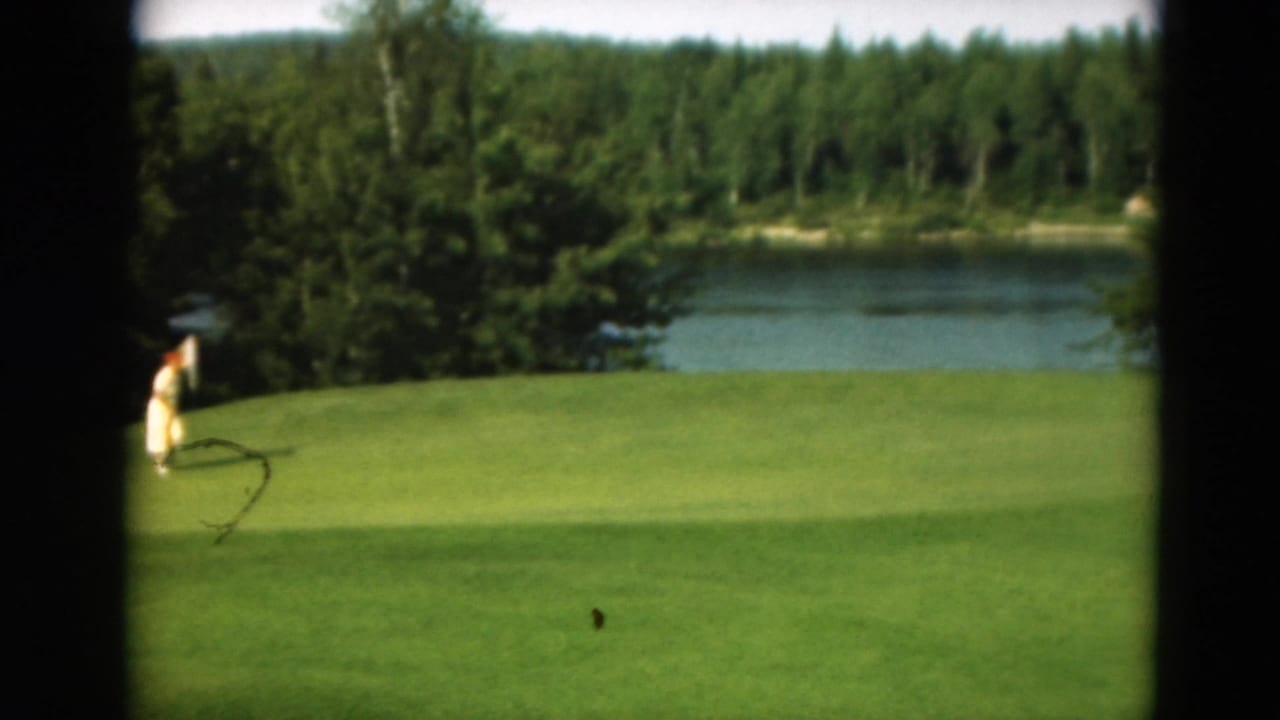 1975 Duluth 8mm frame with a distant woman in a light dress approaching a golf flag on a green overlooking lake water and trees.