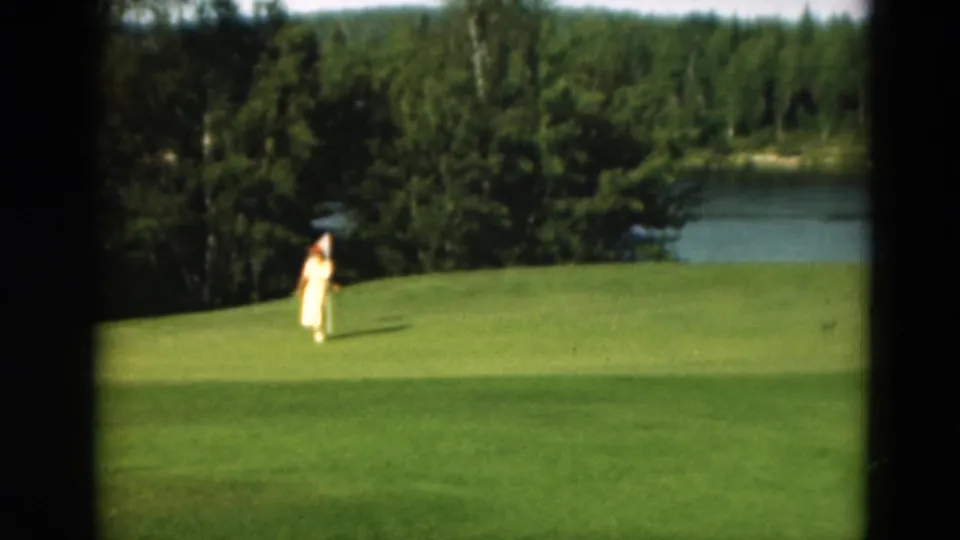 1975 Duluth 8mm frame showing a woman on a green, with clipped grass in front and a wooded lakeshore behind her.