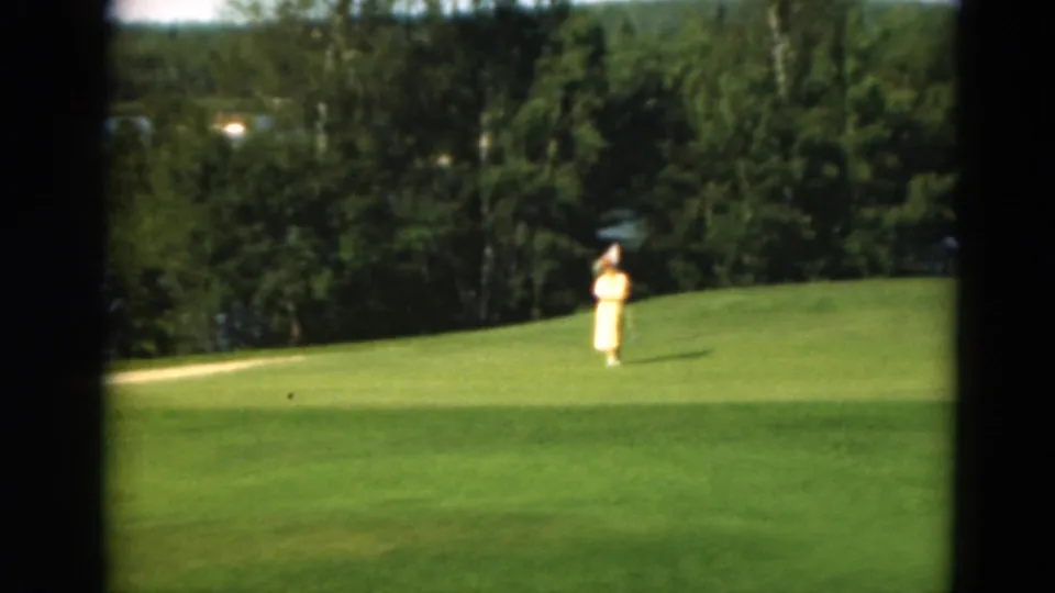 1975 Duluth 8mm frame with a distant woman paused on a golf green, backed by dense trees, lake water and a pale path or sand area.