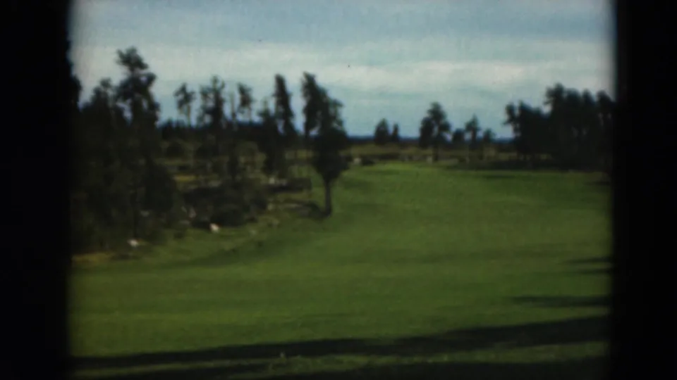 1970s 8mm view of a Minnesota golf fairway, framed by dark edges, with trees and open sky in the distance.