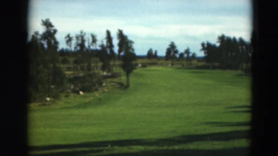 Duluth-area golf-course fairway in 1975 home-movie footage, with trees, hazy sky, and dark edges from the camera position.