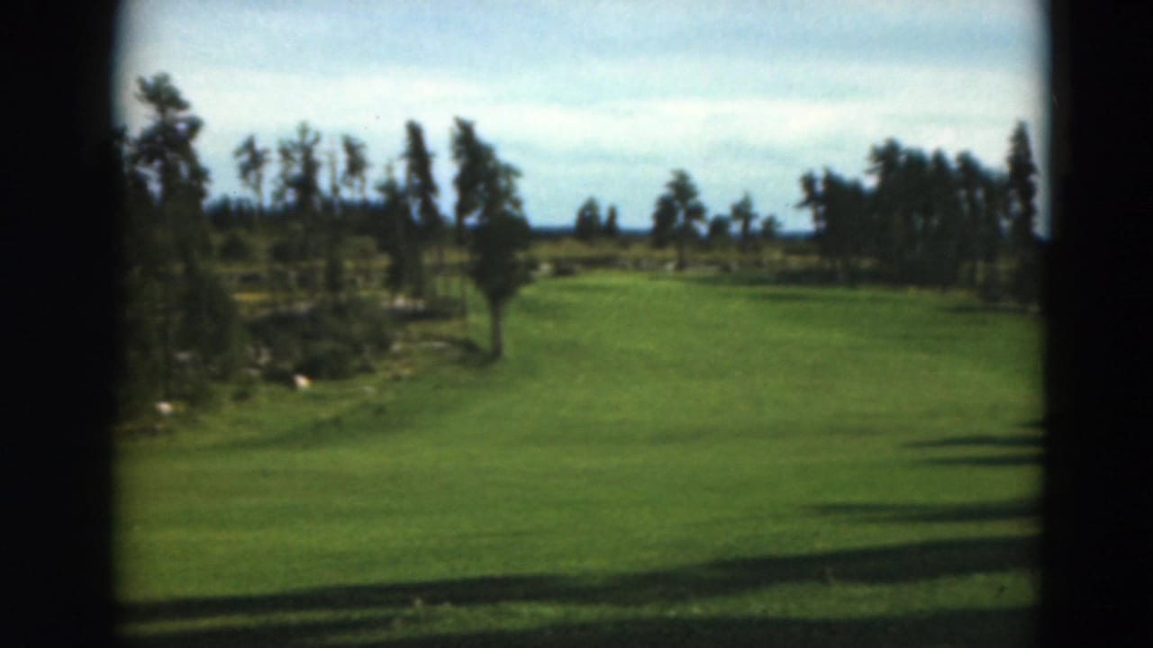 1975 Duluth-area home-movie frame showing an open grassy field, distant tree line, blue sky, and dark film borders; no person is clearly visible in this frame.