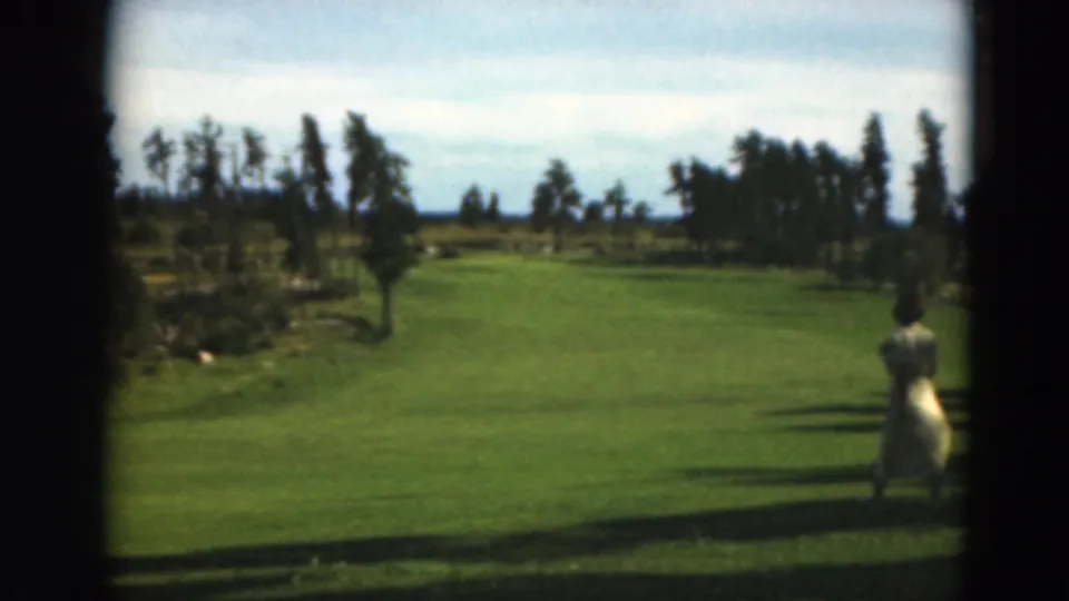 A woman in a white or light dress appears at the right side of a Duluth-area grassy field, walking along the edge of the sunlit lawn in 1975 8mm footage.