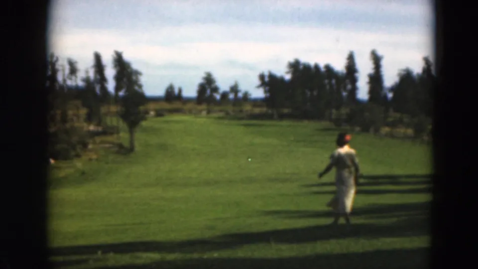 The woman walks across the sunlit grassy field, her light dress and small red head covering standing out against the green lawn and distant trees.