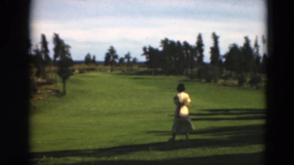 Near the end of the 1975 Duluth-area clip, the woman continues across the open lawn with trees, sky, and long shadows visible around her.