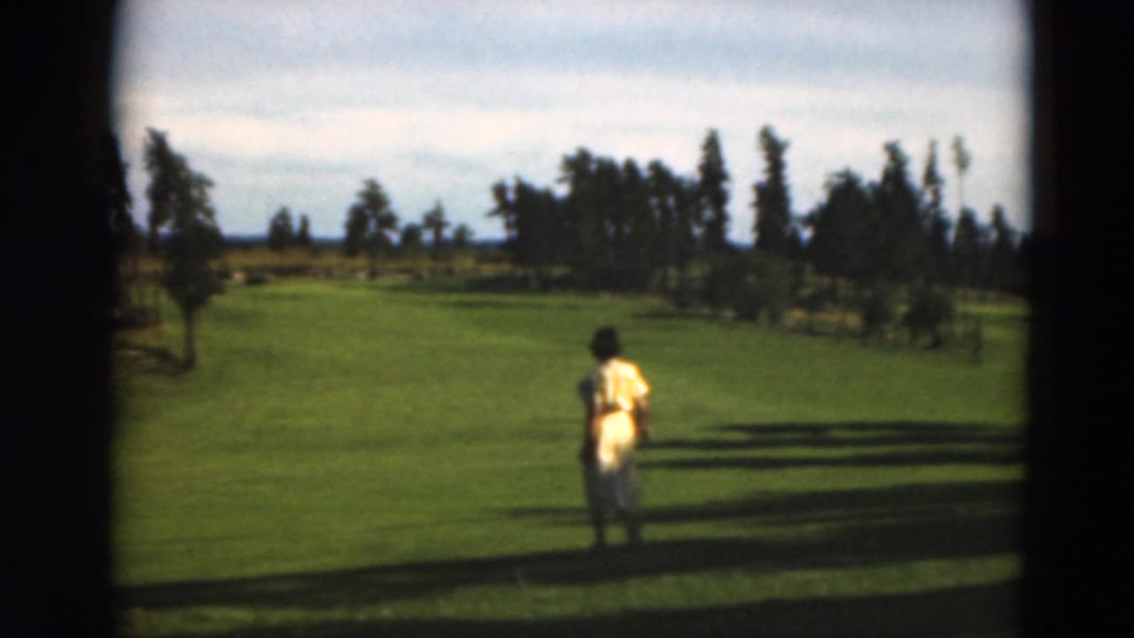 Woman in a yellow dress stands on a green fairway with trees and sky behind her.