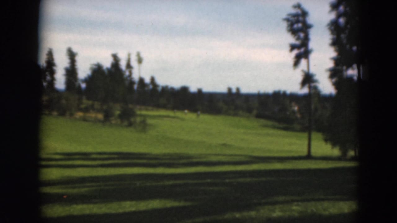Wide 1975 Duluth golf course frame showing a sunlit fairway, tree line, shadows, and dark 8mm scan borders.