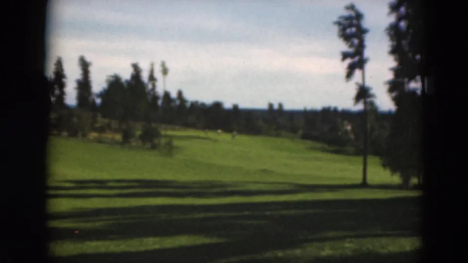 Wide 1975 Duluth golf course frame showing a sunlit fairway, tree line, shadows, and dark 8mm scan borders.