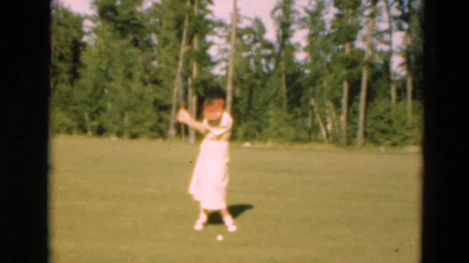A woman in a light dress swings a golf club on a sunlit 1975 Duluth, Minnesota course, with trees behind her.