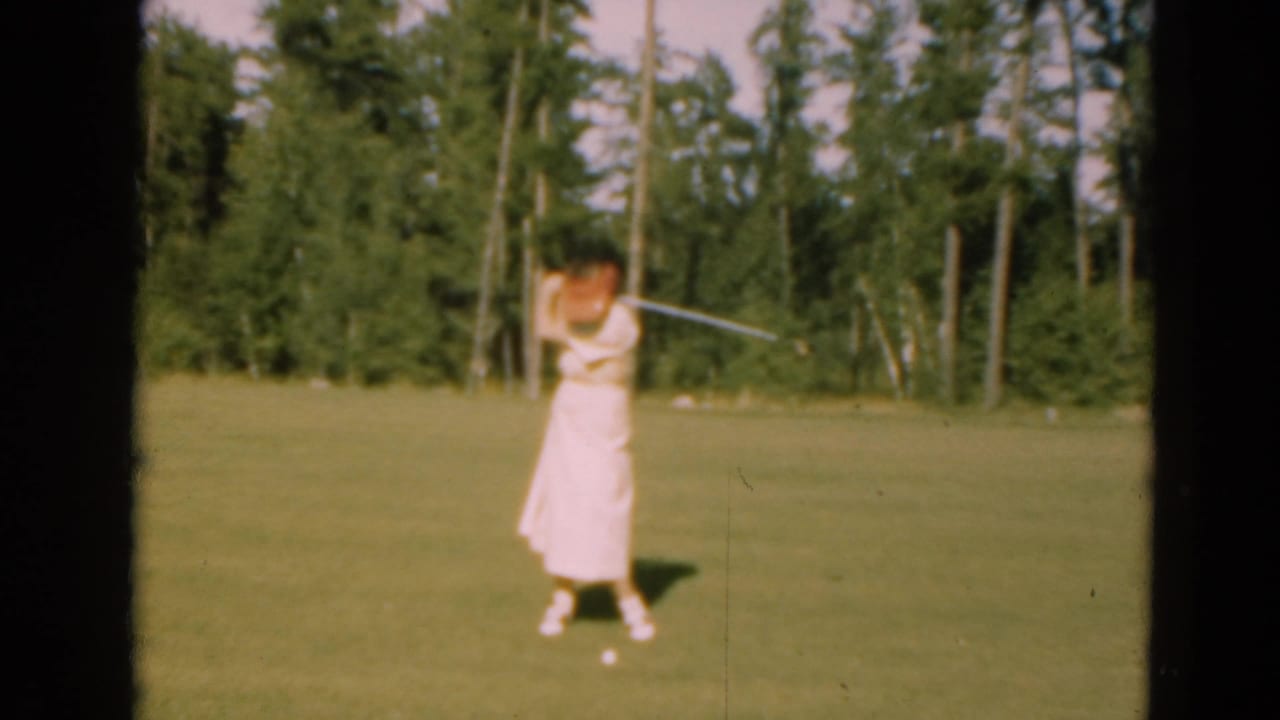 Woman in a light dress swings a golf club on a sunny course bordered by trees.