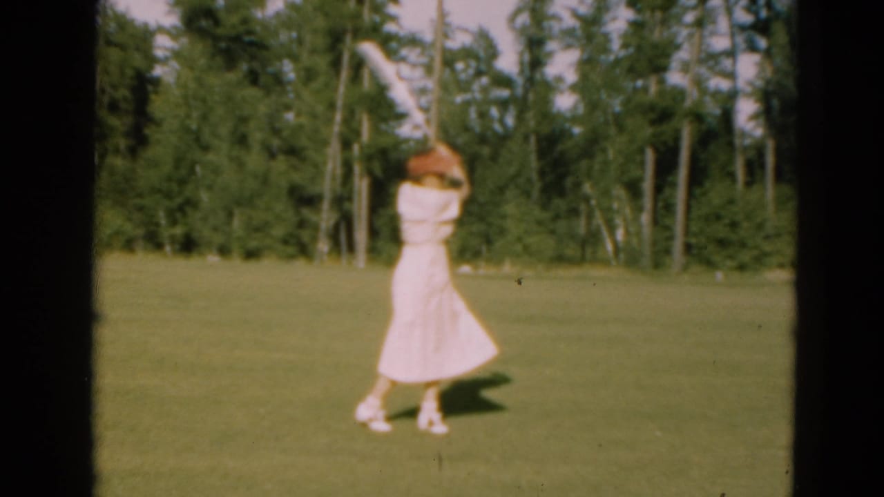 A woman in a pale dress follows through on a golf swing on a Duluth course, with tall trees and open grass visible behind her.