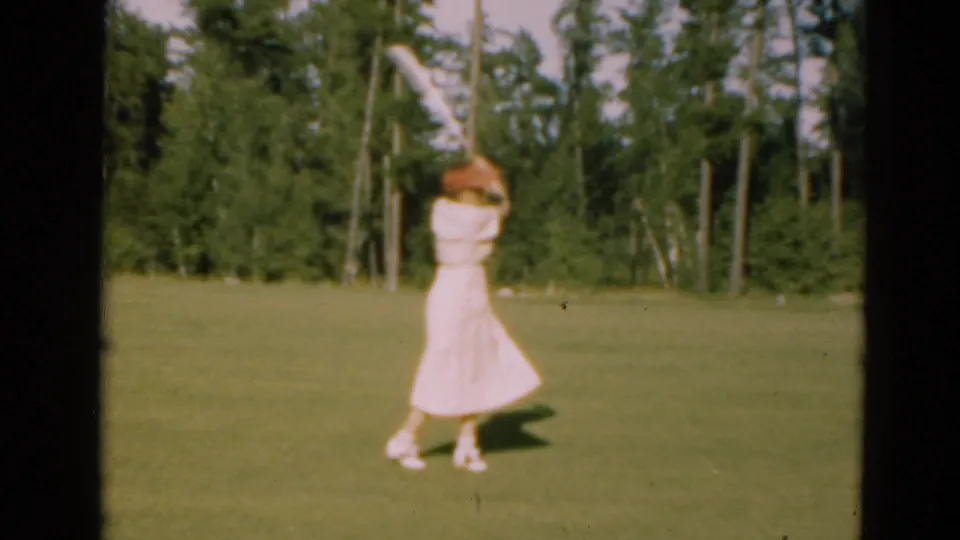 A woman in a pale dress follows through on a golf swing on a Duluth course, with tall trees and open grass visible behind her.