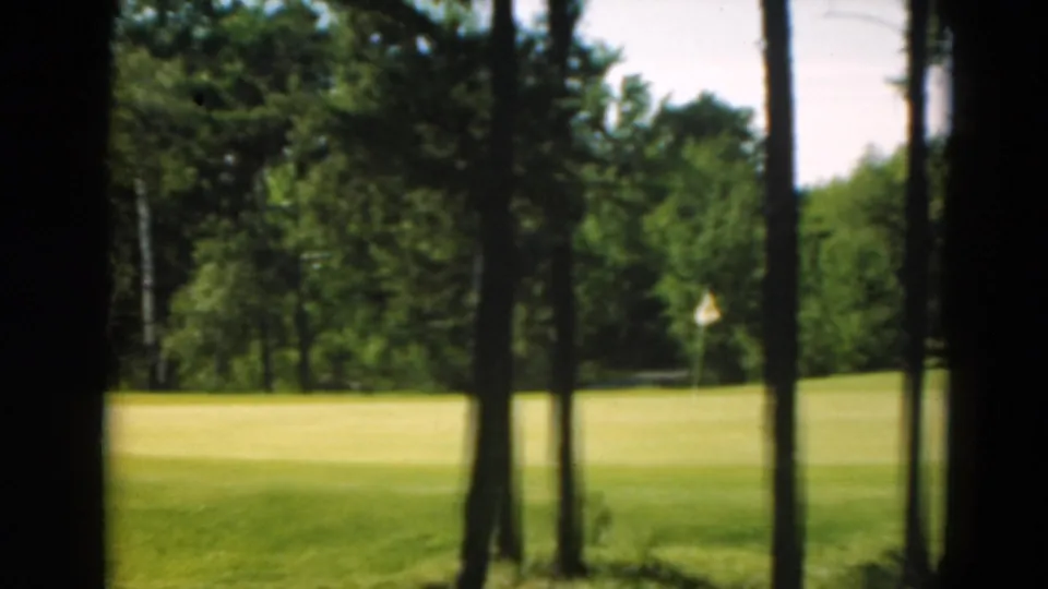 Duluth 1975 home-movie frame showing foreground trees, a bright fairway, and a white golf flag in the distance.