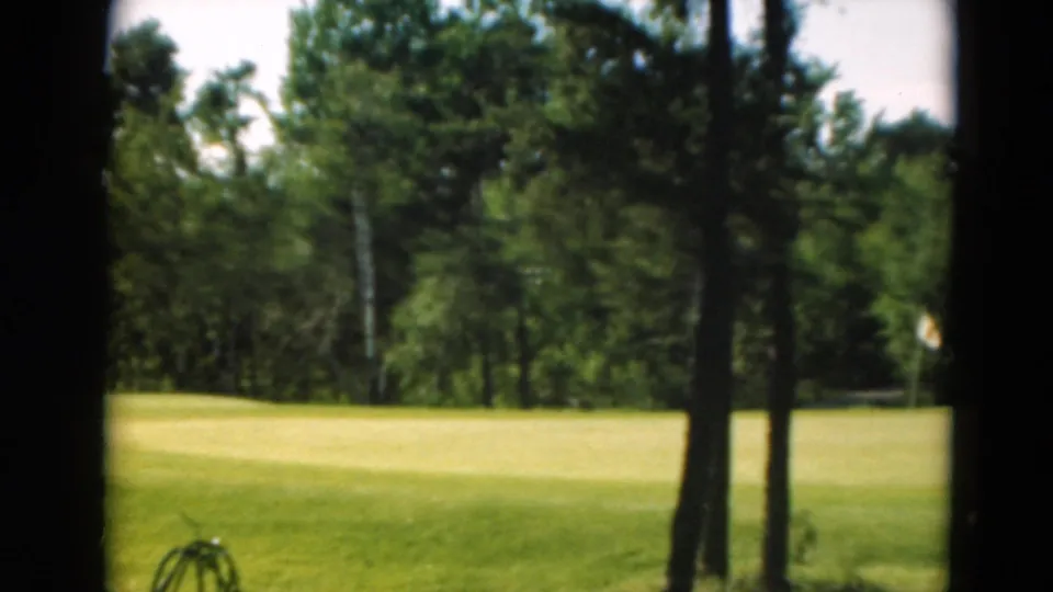 Final 1975 Duluth golf-course frame with trees, bright fairway, wooded background, and a partial bicycle at the lower edge.