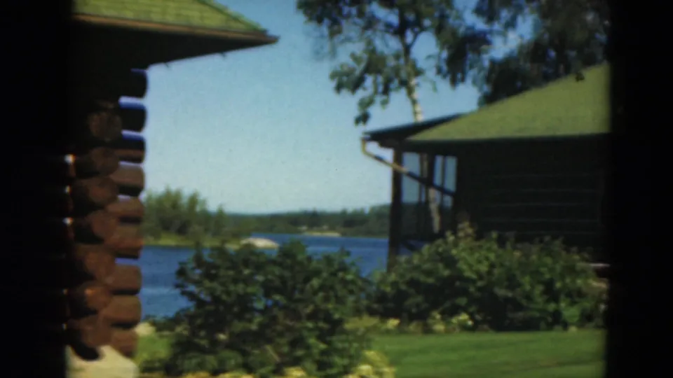 Duluth-area 1975 8mm frame showing log cabin architecture near a blue lake and landscaped greenery.
