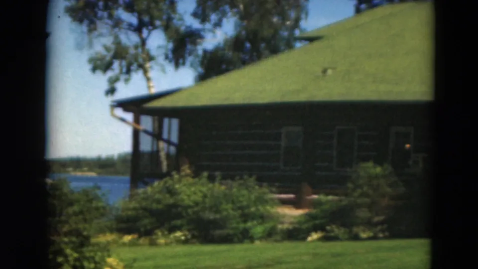 1975 Minnesota lakeside cabin view with green roof, shrubs, lawn, blue water, and distant shoreline.