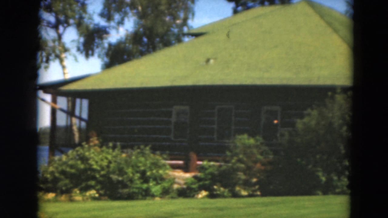 Green-roofed log house behind lawn and shrubs, partly framed by film gate borders.