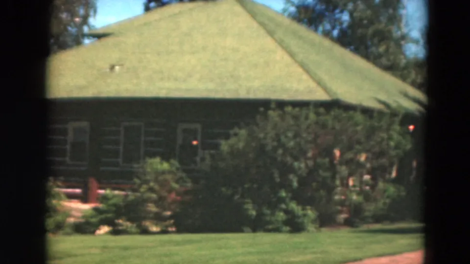 1975 Duluth exterior view of a green-roofed log-style house surrounded by lawn, shrubs, and tall trees.