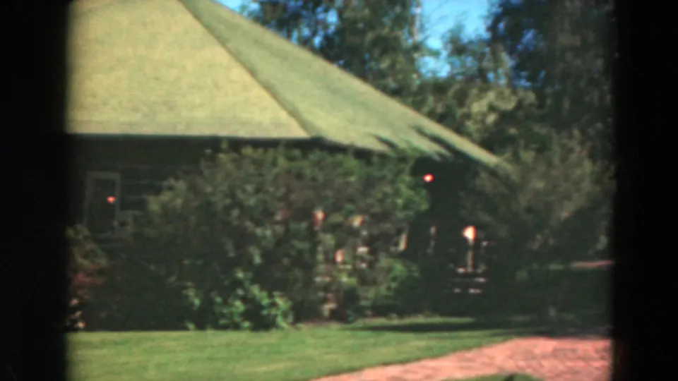 1975 Duluth home-movie frame with the green roof, dense shrubs, lawn, and a reddish path near the rustic house.