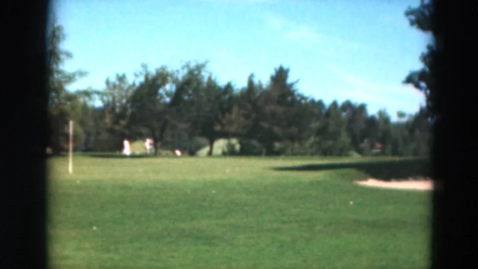 1975 Duluth home-movie view of a golf course with open fairway, flagstick, sand trap, tree line, and distant players.
