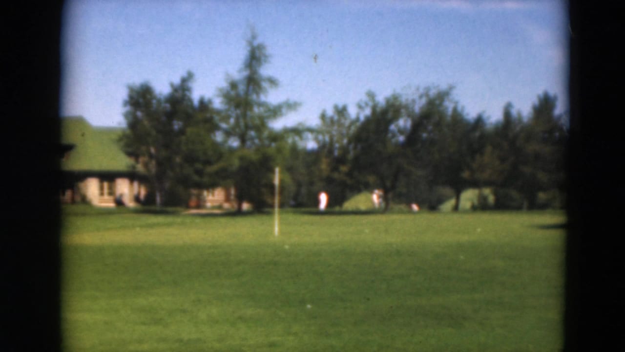 1975 Duluth home-movie frame showing a distant green-roofed lodge across a manicured golf course with trees and small figures.