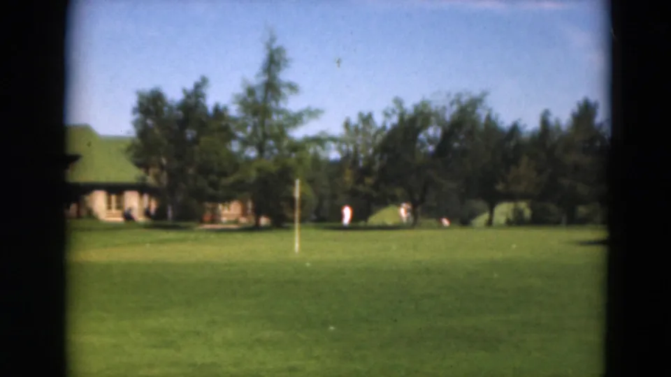 1975 Duluth home-movie frame showing a distant green-roofed lodge across a manicured golf course with trees and small figures.