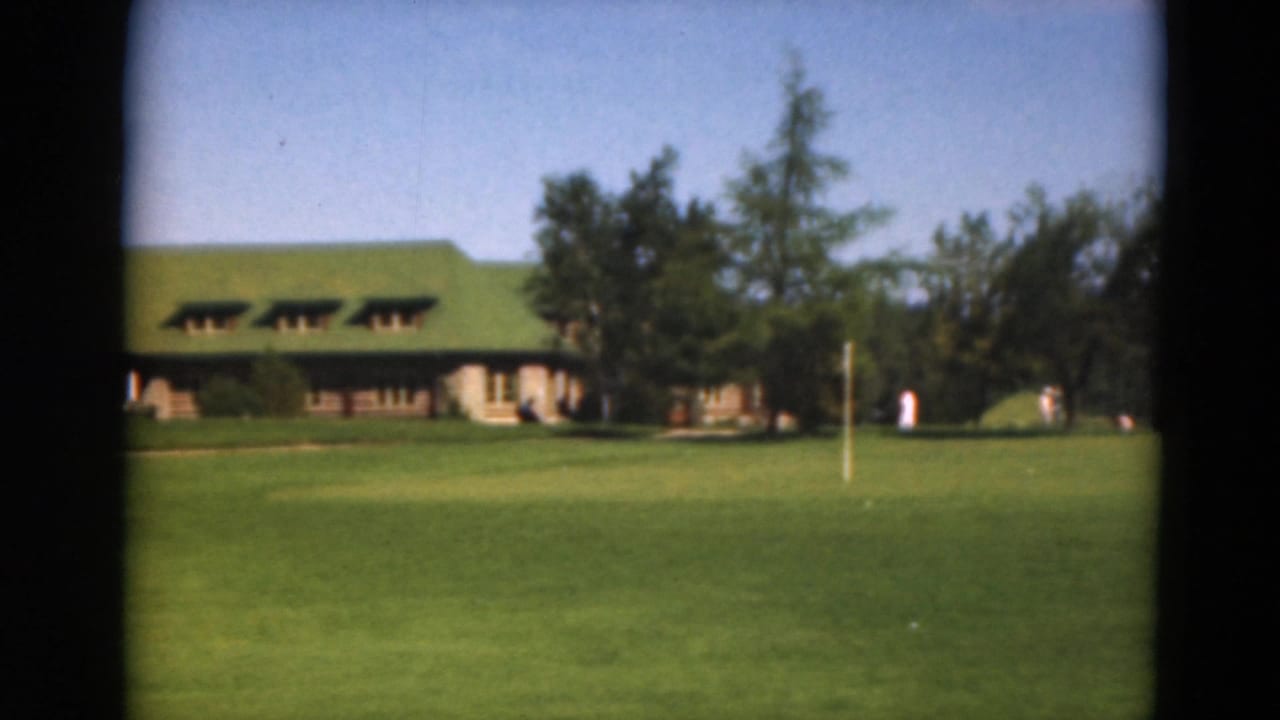 Golf-course lawn with green-roofed building and distant people near trees.
