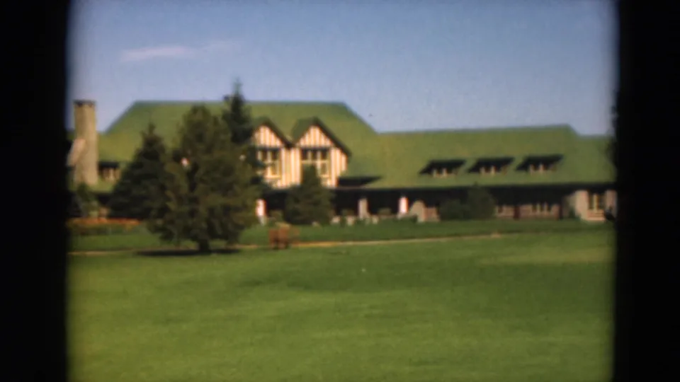 1975 Duluth home-movie frame showing a green-roofed golf-course building with Tudor-style details, evergreens, fairway, and distant figures.