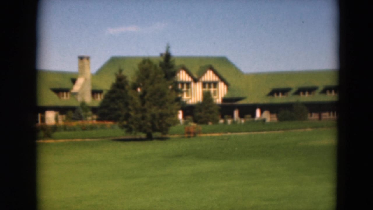 1975 Duluth 8mm frame showing a lodge-style building with a green roof, stone chimney, striped facade, lawn, and evergreens.