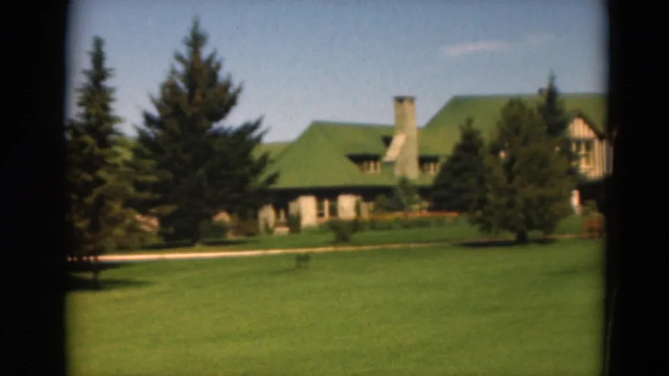 1975 Duluth 8mm frame with evergreens in the foreground, a green-roofed lodge-like building behind them, and open grass in front.