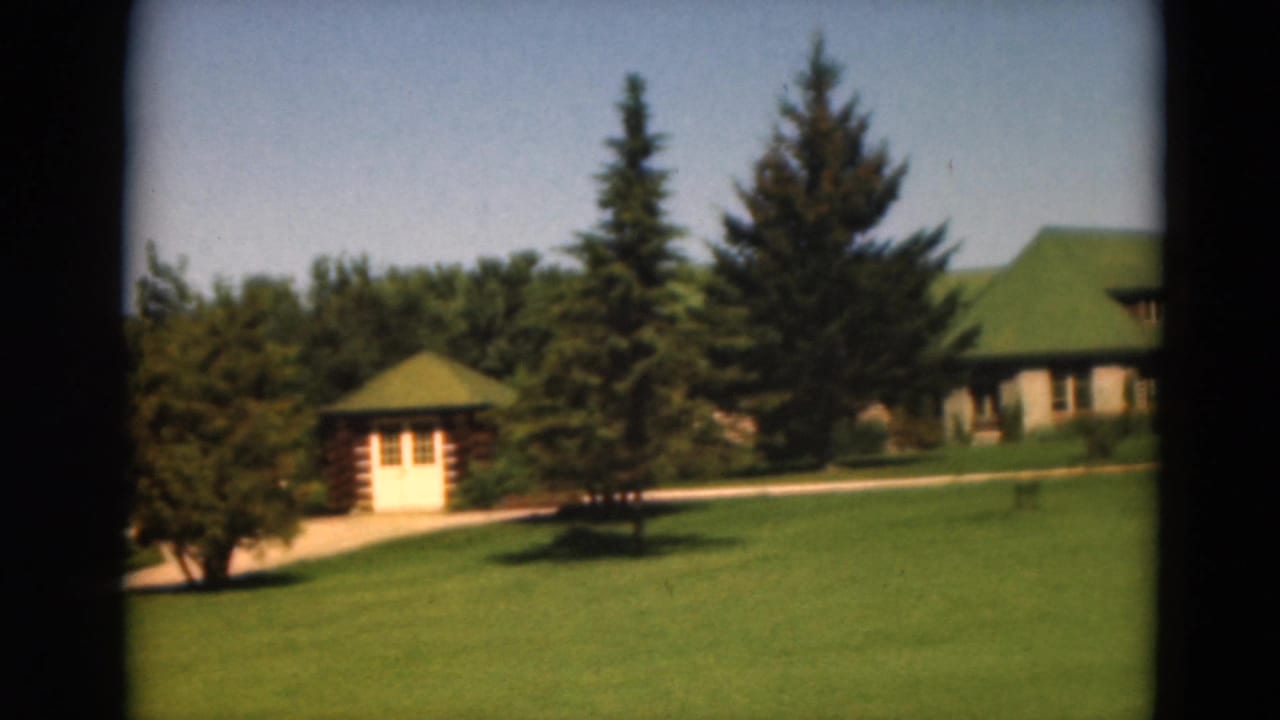 Green lawn with path, small log cabin, trees, and a larger green-roofed building behind it.