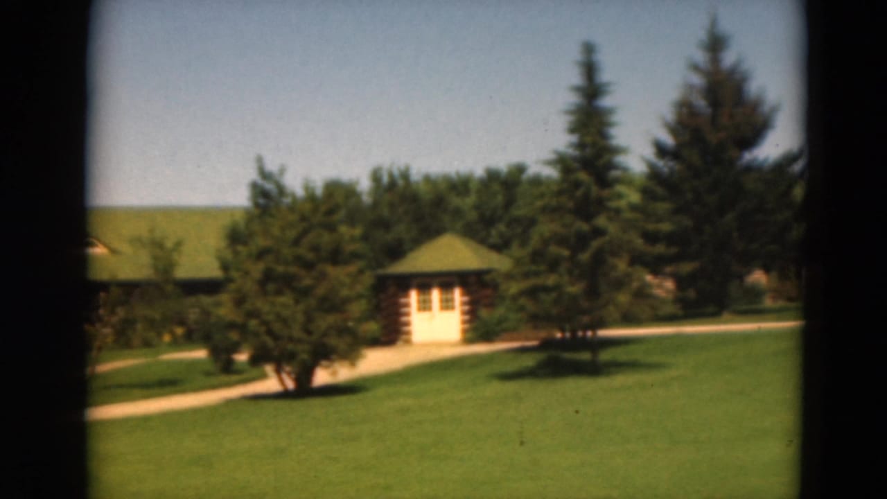 1975 Duluth home-movie frame of a landscaped lawn with path, small cabin, mature trees, and green-roofed structures.