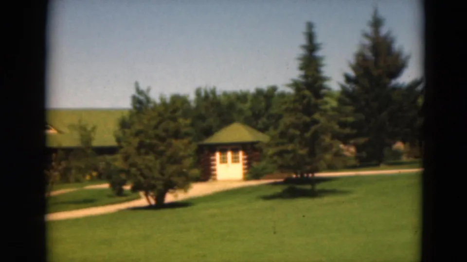 1975 Duluth home-movie frame of a landscaped lawn with path, small cabin, mature trees, and green-roofed structures.