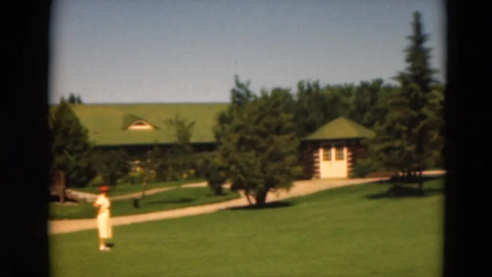 1975 Duluth frame with a distant person in light clothing and red hat crossing a lawn near a path, cabin, and green-roofed building.