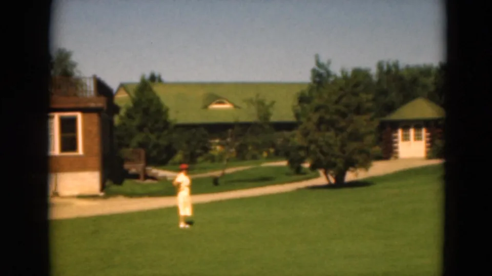 1975 Duluth 8mm frame showing a person standing or walking on the lawn with path, trees, cabin, and green-roofed buildings behind.