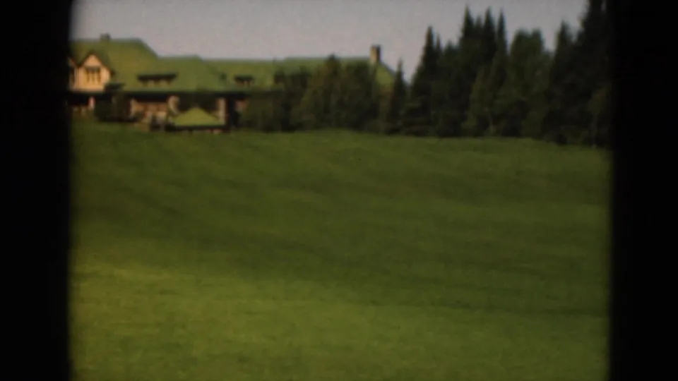 1975 Duluth frame with a broad green lawn leading to a lodge-like building with a green roof and trees.