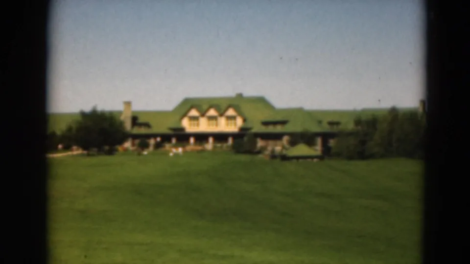 1975 Duluth frame showing a green-roofed house or lodge across the lawn, with porch shade and surrounding trees.