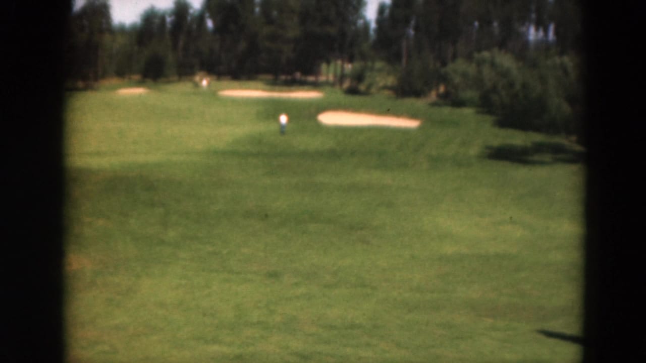 1975 Duluth home-movie frame showing a bright green fairway, sand traps, wooded background, and a distant golfer.