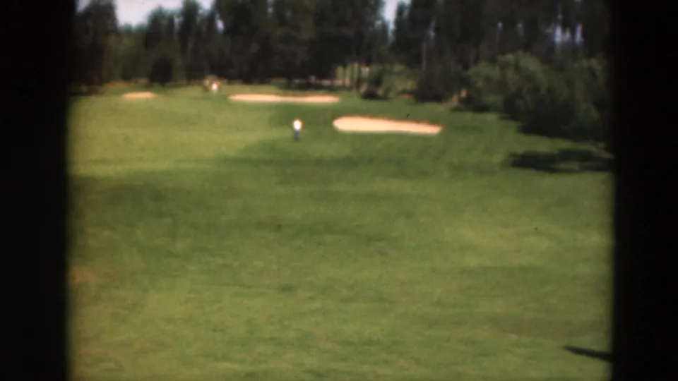 1975 Duluth home-movie frame showing a bright green fairway, sand traps, wooded background, and a distant golfer.