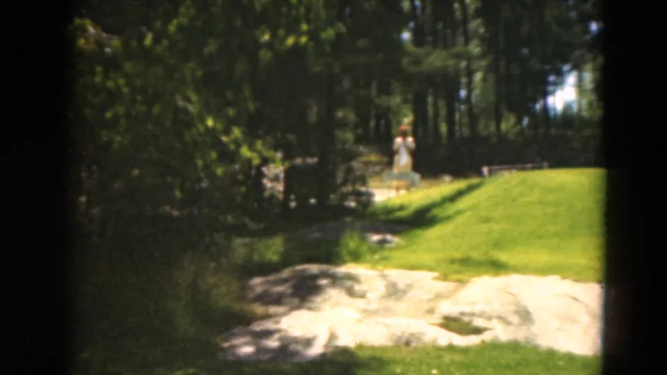 1975 Duluth golf course view with a bright sand bunker, trimmed green grass, dense trees, and a distant person in light clothing near the woods.