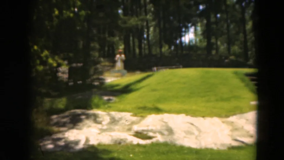 1975 Duluth 8mm frame showing a sand bunker, bright green course surface, wooded background, and a small distant figure near the tree line.