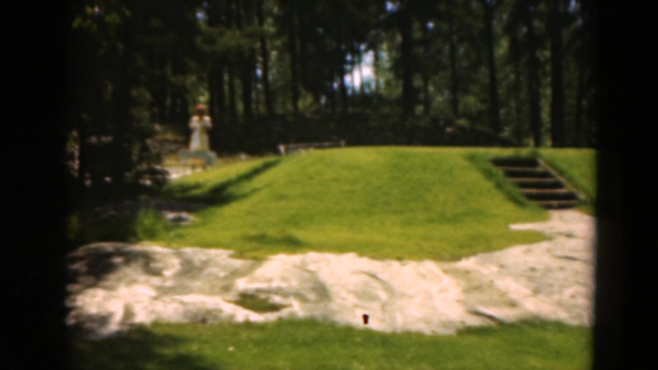 Golf green with sand trap, wooded background, steps, and a small pale figure near the trees.