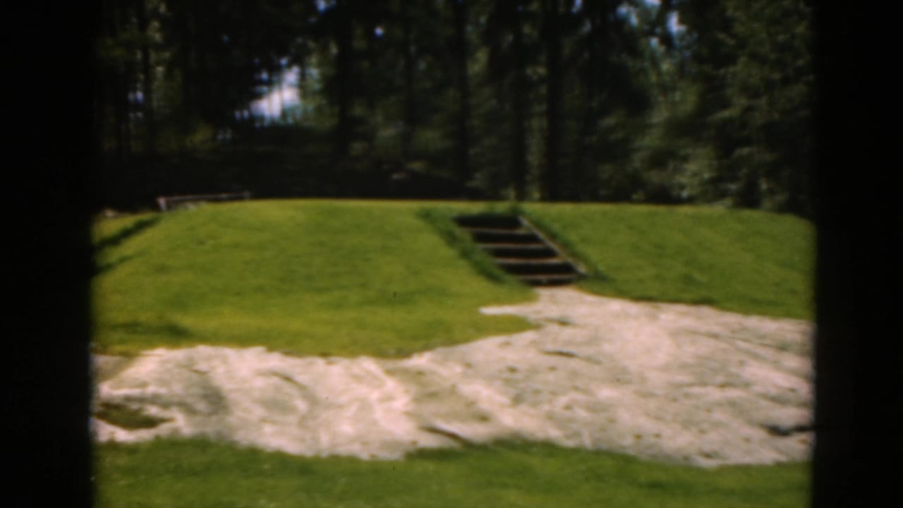 1975 Duluth 8mm view of a grassy golf green, pale sand area, wooden steps, and dark evergreen background.