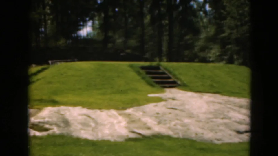 1975 Duluth 8mm view of a grassy golf green, pale sand area, wooden steps, and dark evergreen background.