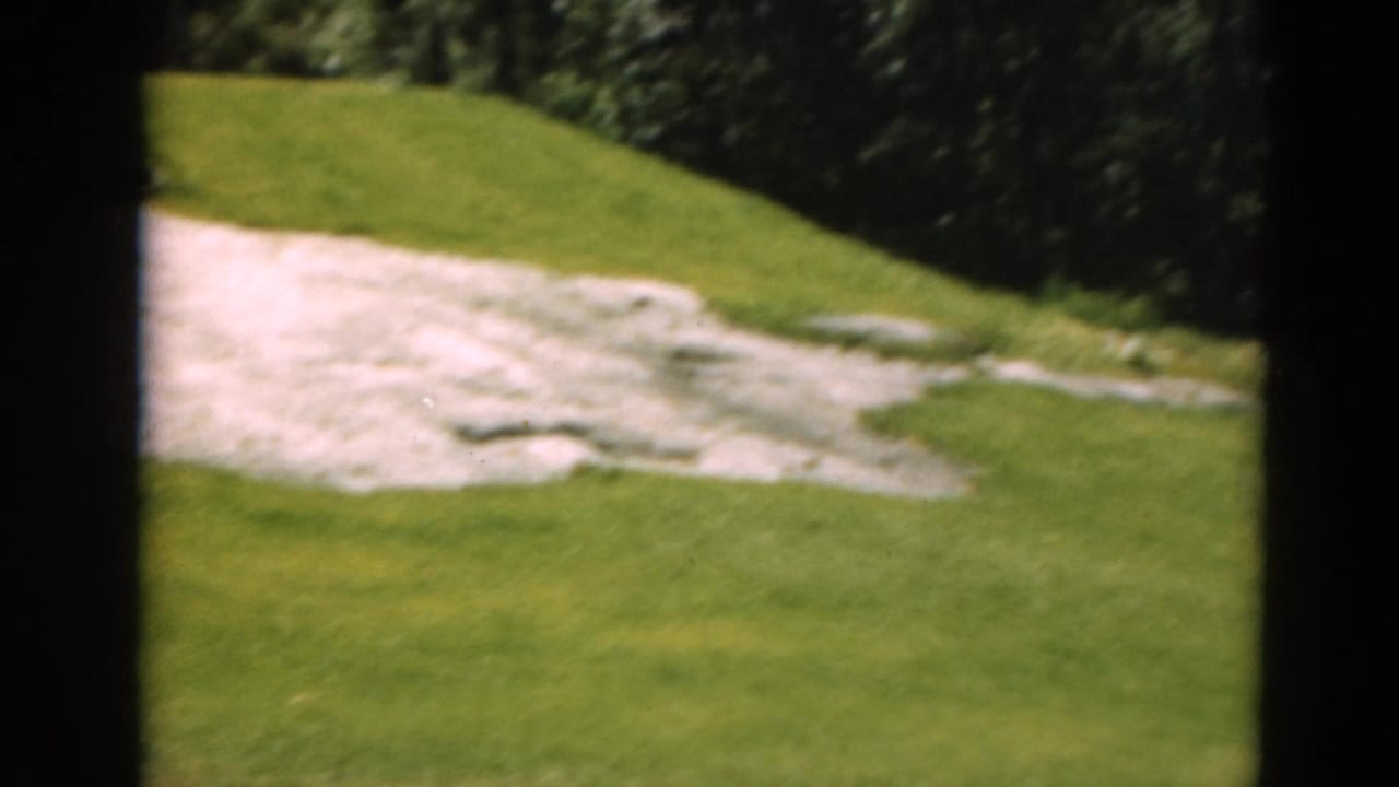 Duluth golf course view on 8mm film with a pale bunker set into green grass near a wooded edge.