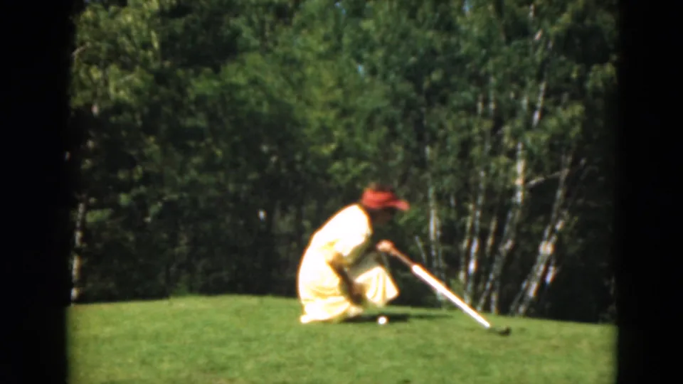 A golfer in a yellow dress and red cap crouches with a putter on a Duluth golf green in 1975 home-movie footage.