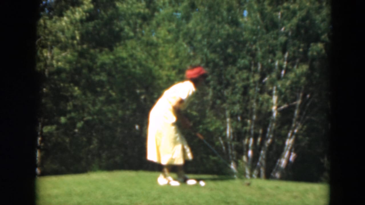 Woman in a yellow dress and red hat bends over a golf ball on a green.