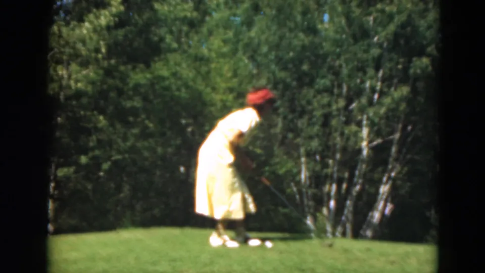In Duluth, Minnesota in 1975, a woman in a yellow dress and red hat leans toward a golf ball on a sunlit green.