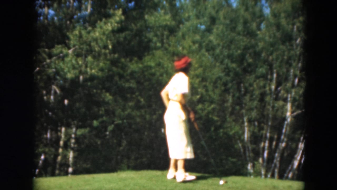 A woman golfer in a yellow dress and red hat stands on a Duluth green with trees behind her in 1975 home-movie footage.